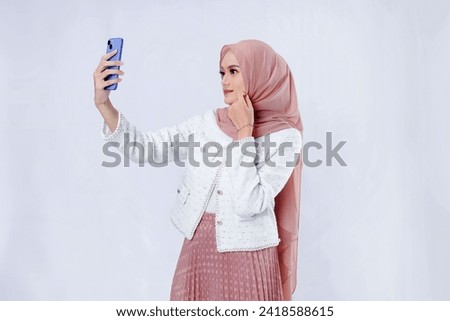 Asian hijab woman taking a selfie using a cellphone while smiling on a white background