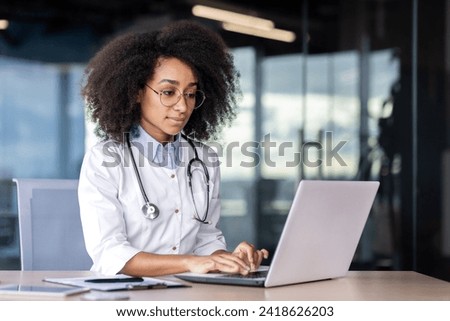African american female therapist dressed in uniform typing on laptop while sitting at workplace in hospital. Pretty physician using modern device and internet for helping and supporting patients.