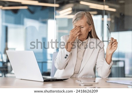 Tired senior woman dressed in office suit sitting at desk with laptop and touching forehead while having headache. Female director taking off glasses to let eyes rest during working at computer. Royalty-Free Stock Photo #2418626209