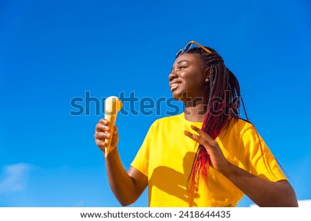 African woman with braided hair eating an ice cream under the sun with blue sky on the background