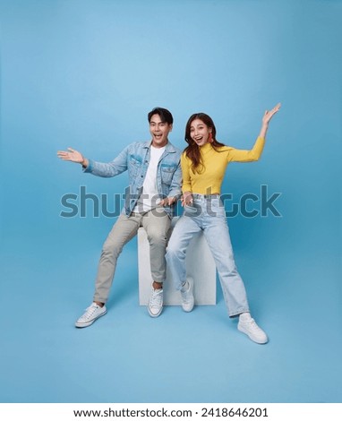 Happy Asian couple sitting back to back on white cube chair and presenting or showing open hand palm with for product isolated on blue studio background.