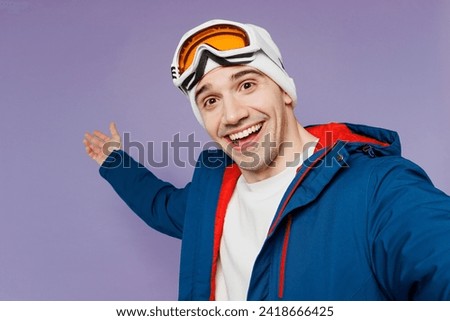 Close up skier man wear blue windbreaker jacket ski goggles mask hat do selfie shot mobile cell phone point aside spend extreme weekend winter season in mountains isolated on plain purple background