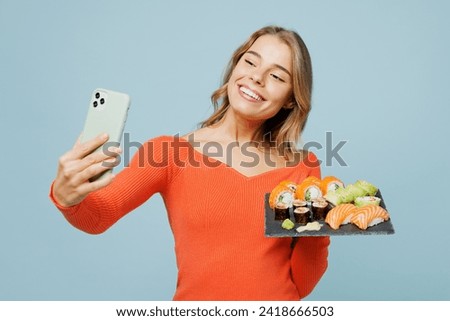 Young woman wear orange casual clothes do selfie shot mobile cell phone hold eat raw fresh philadelphia sushi roll served on black plate Japanese food isolated on plain blue background studio portrait