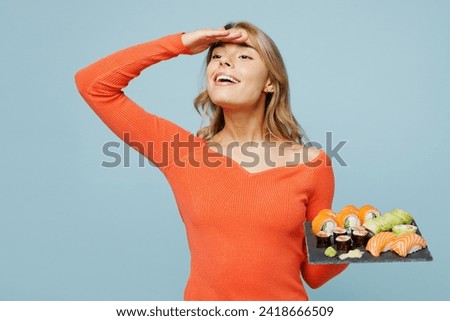 Young happy woman wear orange casual clothes hold hand at forehead look far away distance eat raw fresh sushi roll served on black plate Japanese food isolated on plain blue background studio portrait