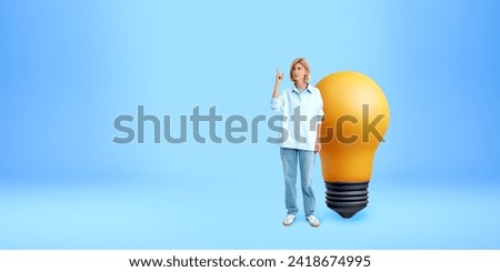 Woman finger pointing up, full length standing near large light bulb on empty copy space blue background. Concept of inspiration, start up, creativity, knowledge and idea