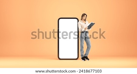 Happy woman full length reading a paper document in hands, standing near large mockup phone screen. Concept of electronic documents, files storage, database and mobile app