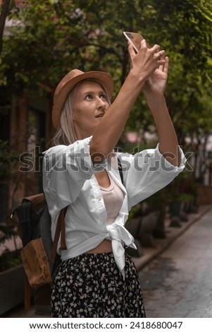 Stylish young blonde woman in a hat, white shirt with a backpack takes pictures on a beautiful street in the city on a mobile phone. Young female traveler. Vertical photo