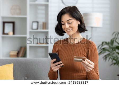 Smiling young woman sitting on sofa at home, holding credit card and using mobile phone. Close-up photo.