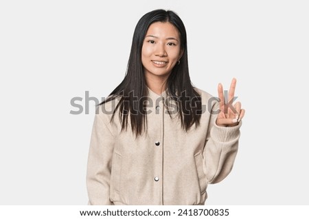 Young Chinese woman in studio setting showing victory sign and smiling broadly.