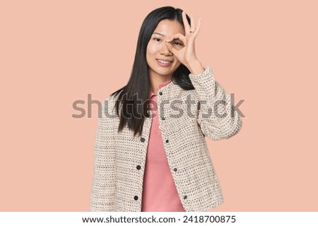 Young Chinese woman in studio setting excited keeping ok gesture on eye.
