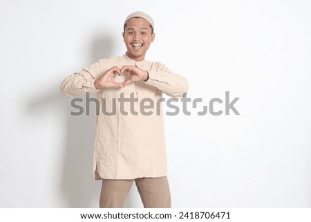 Portrait of excited Asian muslim man in koko shirt with skullcap speaks own feelings, makes heart gesture over chest, expresses sympathy and love, smiles positively. Isolated image on white background