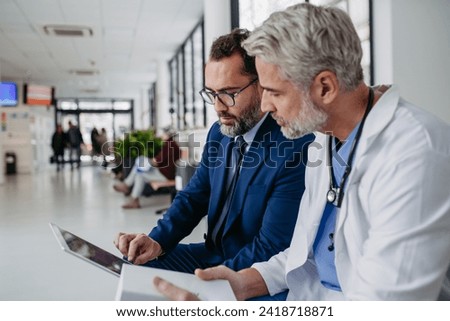 Pharmaceutical sales representative talking with doctor in medical building, presenting new medication on tablet. Hospital director, manager in modern clinic with surgeon. Royalty-Free Stock Photo #2418718871