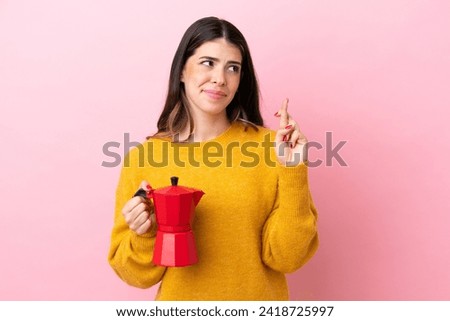 Young Italian woman holding a coffee maker isolated on pink background with fingers crossing and wishing the best
