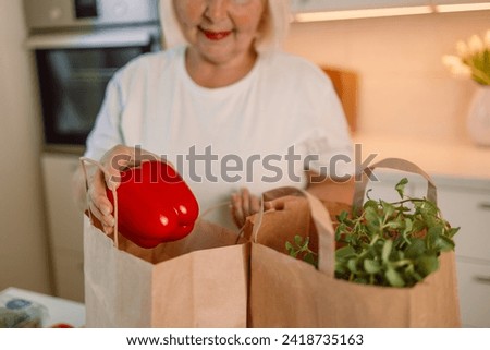 Happy lady unpacking groceries at home. Portrait of cheerful beautiful woman at kitchen table counter with bags full of apples, oranges, peppers, tomatoes, zucchinis, parsley from food market. High Royalty-Free Stock Photo #2418735163