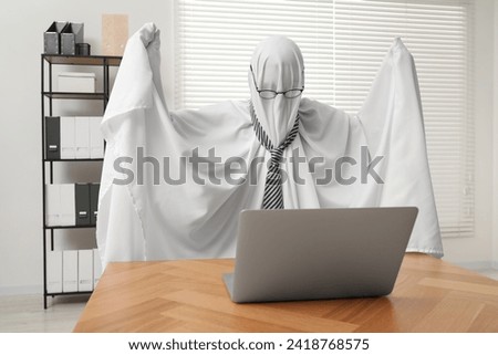 Overworked ghost. Man covered with white sheet using laptop at wooden table in office Royalty-Free Stock Photo #2418768575