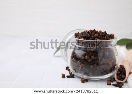 Aromatic cloves in glass jar and scoop on white tiled table, closeup. Space for text