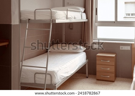 A clean and minimalistic hostel room with a bunk bed, white bedding, and a small wooden bedside table, ideal for budget travelers seeking affordable accommodation. Cheap hostels, save money. Royalty-Free Stock Photo #2418782867
