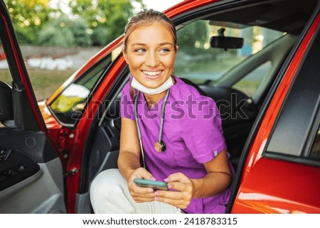 A smiling young nurse dressed in her scrubs uniform sitting in her car holding her mobile phone, taking a break from work. Royalty-Free Stock Photo #2418783315