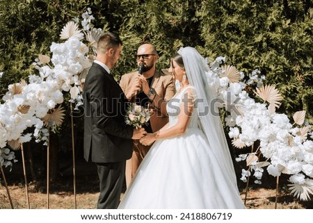 Wedding ceremony in nature. The bride and groom near the flower arch. Master of ceremonies in dark glasses at a wedding during a performance against the background of the bride and groom.