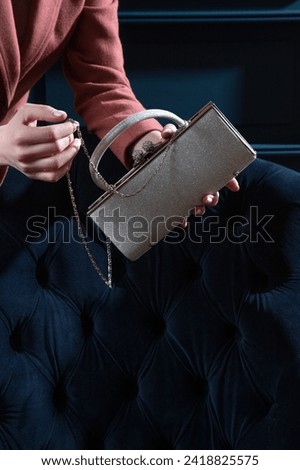 Silver metallic sparkling purse held by woman hands on a blue velour armchair background. Creative shiny woman hand bag studio photography. Royalty-Free Stock Photo #2418825575