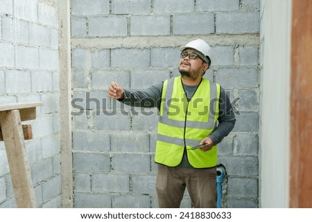 Asian male construction engineer, wearing full clothing, safety helmet, reflective vest, holding laptop in hand, checking work site for completeness, following construction plans. Royalty-Free Stock Photo #2418830633