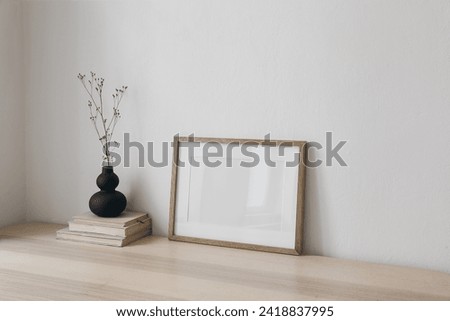 Blank horizontal wooden picture frame mockup. Organic shaped black vase with dry grass on table, desk. Cup of coffee, old books. Working space, home office. Art, poster display, boho beige interior.