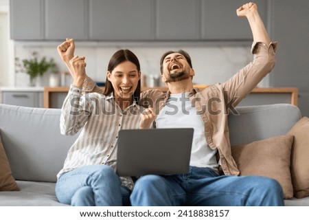 Joyful young caucasian couple seated on sofa, celebrating win with arms raised and laptop in front, resting in bright contemporary living space Royalty-Free Stock Photo #2418838157