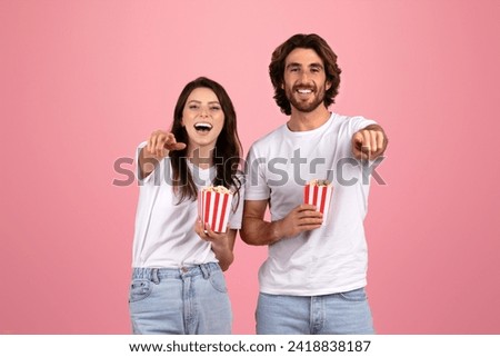 Cheerful european young couple in white t-shirts and jeans, laughing and pointing towards the camera, each holding a small striped popcorn box, on a soft pink background, studio