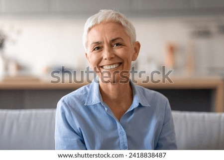Portrait of beautiful senior woman with short gray hair sitting on sofa indoor and smiling looking at camera. Attractive lady in casual blue shirt relaxing at home, enjoying comfort Royalty-Free Stock Photo #2418838487