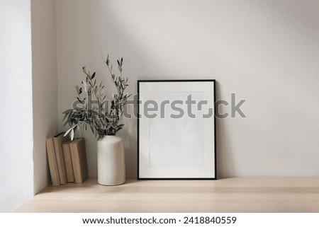 Elegant Mediterranean working space. Books, empty vertical black picture frame mockup. White wall background. Wooden desk, table. Vase with olive branches, home office concept. Scandinavian interior 