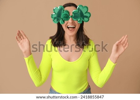 Beautiful young woman with decorative glasses in shape of clover on brown background. St. Patrick's Day celebration