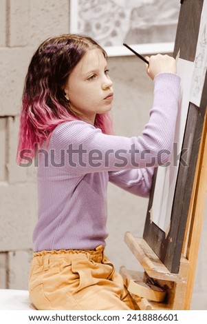 11 years old girl craftswoman are painting on canvas in studio standing in front of easel. Portrait of a girl painting during an art class