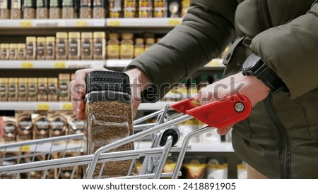 Close-up of a shopping trolley in a coffee department and a buyer putting a jar of instant coffee into it Royalty-Free Stock Photo #2418891905