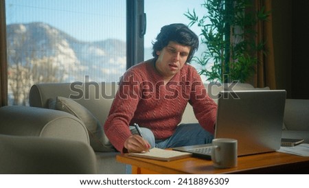 Young college student working from home using with laptop while sitting on sofa in living room at home. Focused millennial male looking at laptop, taking note on notebook