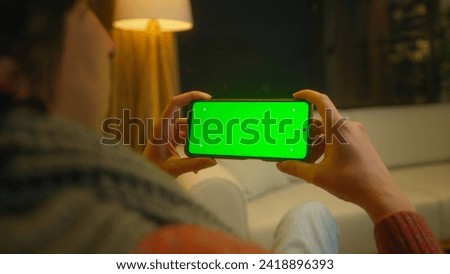 Young man watching video on his smartphone with green screen mock up display, which he holds horizontally. Male sitting on sofa relaxing at home. Close-up over the shoulder shot