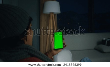 Young man using, tapping on screen smartphone with green mock-up screen. Male sit on armchair, relaxing at home. Close-up over the shoulder shot