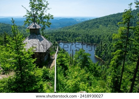 The photo shows a wooden chapel perched on a rocky outcrop surrounded by dense forest above a mountain lake. Taken at the Rachel Chapel in the Bavarian Forest National Park, Germany. Royalty-Free Stock Photo #2418930027