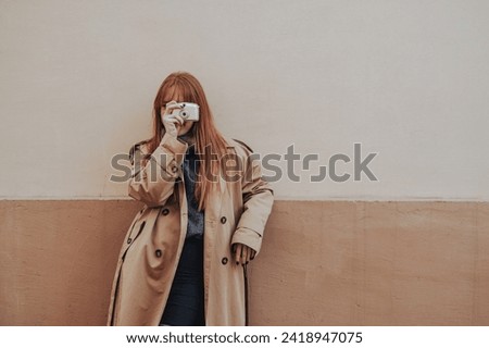 A fashionable young woman is taking pictures with her camera while leaning on a rustic wall on a city street downtown. A young lady in a coat is photographing precious moments on street. Street photo