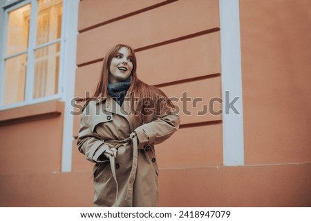 A stylish young woman in warm outfit is adjusting her coat in cold weather in rustic part of the town while looking away. Fashion photo of a young woman posing with her coat on a street. Copy space.