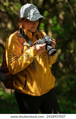 Beautiful Latin American woman walking with a red backpack and blue cap reviewing the photos on her camera in the middle of a forest during a sunny day