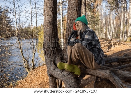 Pensive smiling midle-aged woman sitting on pine tree root, enjoying sunny warm autumn day, thinking of life, dreaming, listening to silence. Recreation, relaxation in nature, pleasant time spending Royalty-Free Stock Photo #2418957197