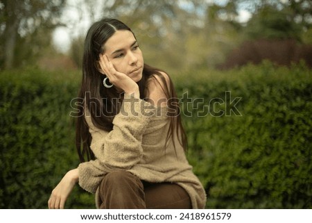 A young Caucasian woman sits outdoors, chin resting on hand against a blurred plant background, directing her gaze towards copy space. versatile visual with emphasis on nature and customizable content