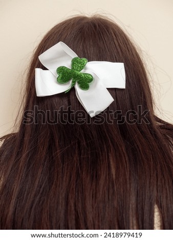 White bow with green shamrock attached on long brown hair of a girl. Light color background. Dressing up for special Irish community day. Popular symbol in Ireland. Celebrating Saint Patrick day