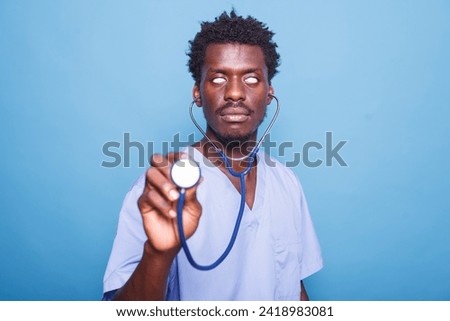 In studio, playful male nurse is clutching a stethoscope and rolling his eyes. Against blue background, African American medical assistant holds a cardiology instrument for a heartbeat examination. Royalty-Free Stock Photo #2418983081