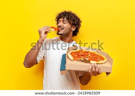 young indian man eating delicious pizza on yellow isolated background, curly guy student holding pizza box and biting a piece of fast food Royalty-Free Stock Photo #2418983509