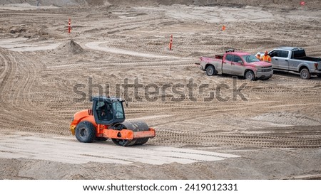 Vibratory soil roller compactor compacting a thickness of sand on a construction site, with two pick-ups and workers Royalty-Free Stock Photo #2419012331
