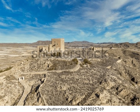 Aerial view of Jumilla medieval castle in Murcia Spain, on a hilltop, imposing irregular shape keep with four floors, crenelated battlements cloudy blue sky background