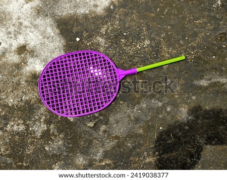 A small children's toy racket made of purple and light green plastic, the handle is broken and missing. Located outside the front of the house.