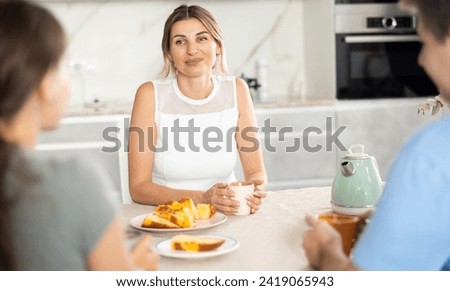 Cheerful young woman receiving guests over cup of tea with freshly baked sweet pie at home, sitting at cozy kitchen table and talking friendly