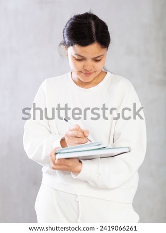 Positive interested young Asian female college student in white casual clothes holding notebooks standing on gray background, making notes. Studying concept..